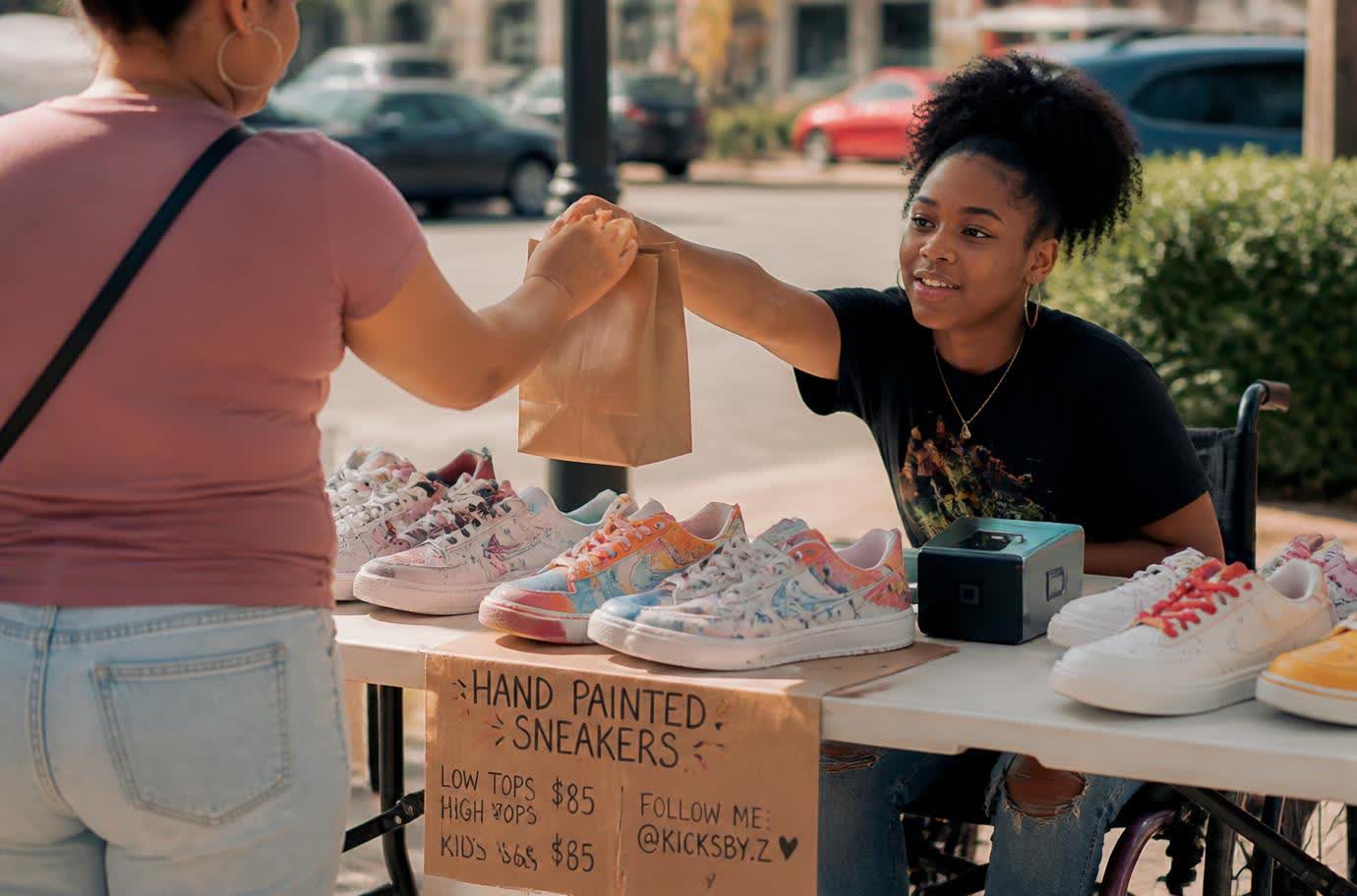 A teenage girl using a wheelchair hands a paper bag to a customer across a market table of custom-painted sneakers, with a card reader beside them.